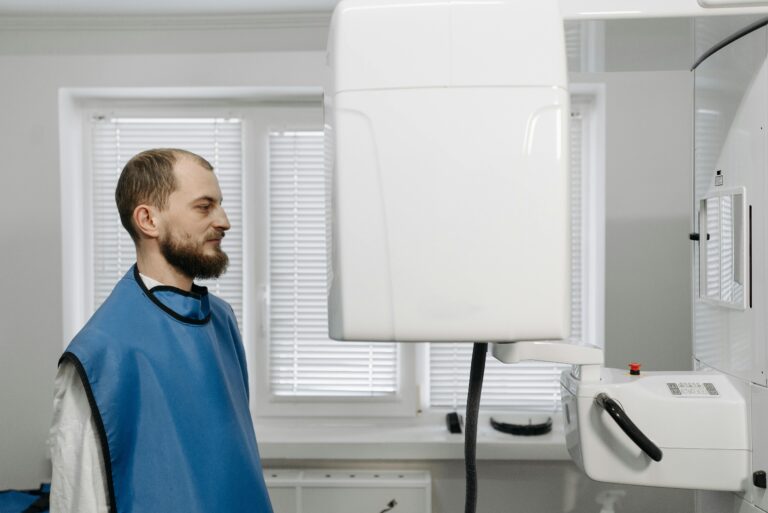 A man in a medical gown receiving a dental x-ray examination in a clinical setting.